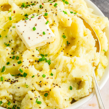 garlic mashed potatoes in a white bowl with a pat of dairy free butter and chives as garnish and a spoon to the right inside the bowl.