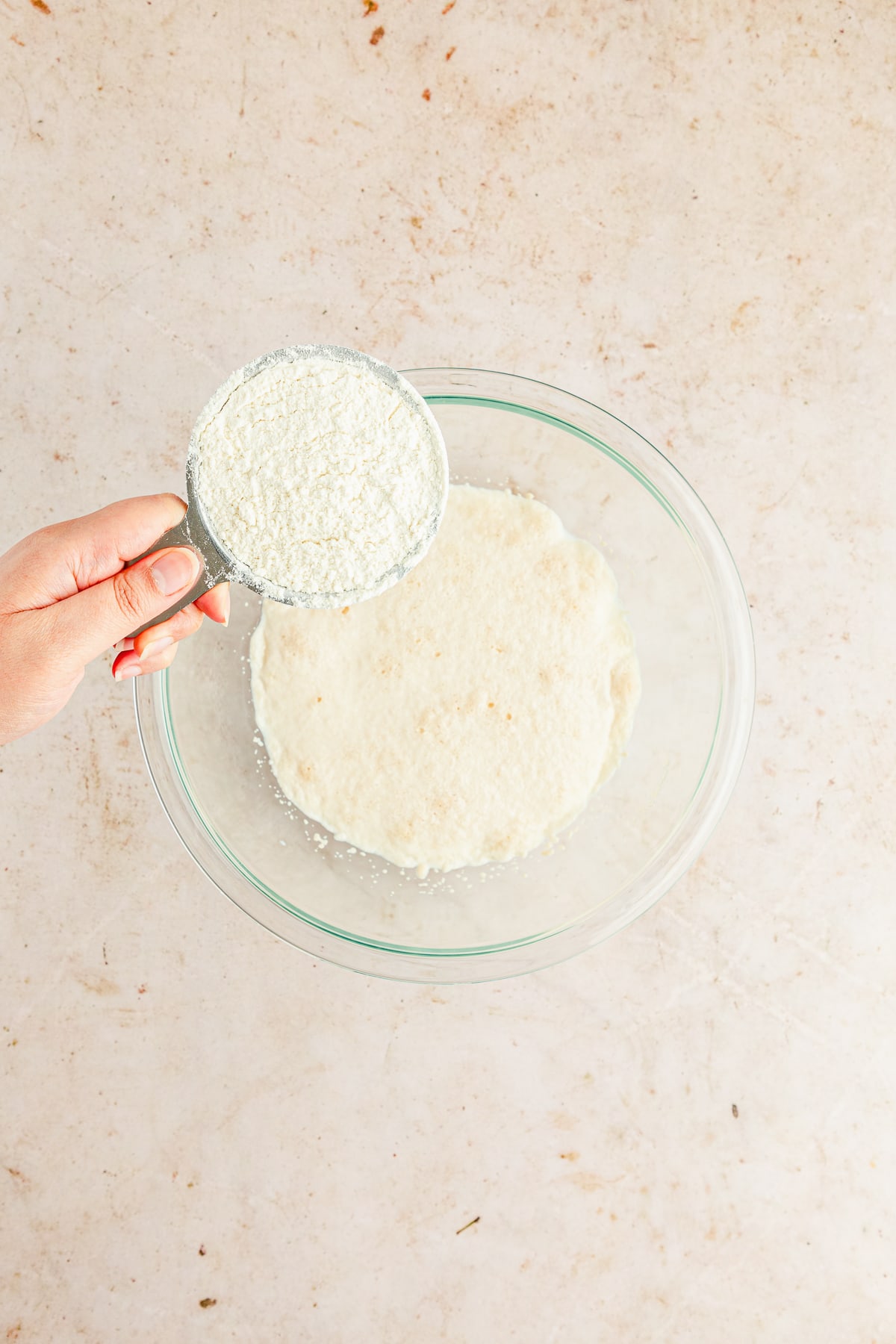 hand holding a cup of flour in a measuring cup over a mixture for dough in a glass bowl.