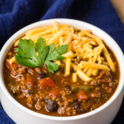 instant pot chili in a white bowl with shredded cheddar cheese and herb garnish on a navy blue cloth napkin.