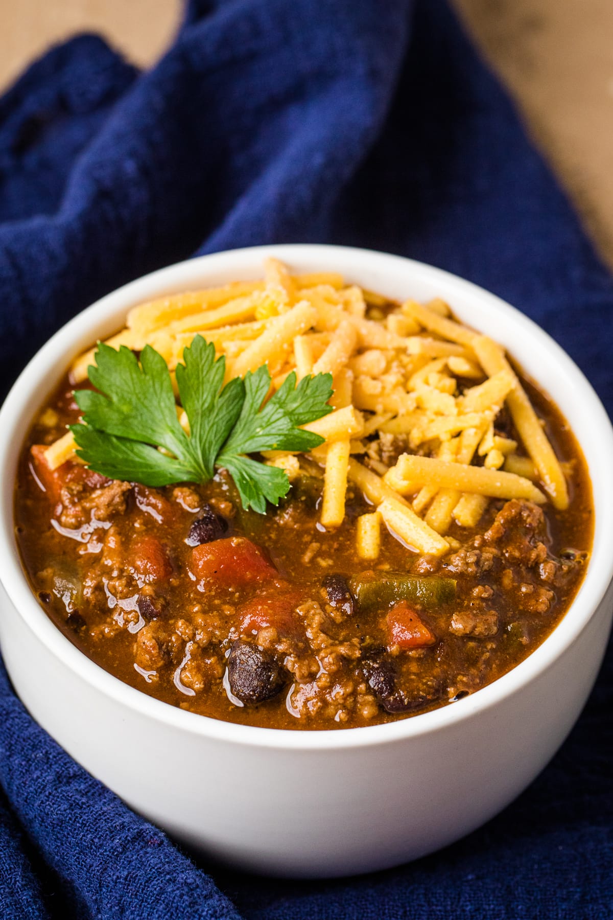 instant pot chili in a white bowl with shredded cheddar cheese and herb garnish on a navy blue cloth napkin.