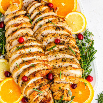 sliced roast turkey breast on a white platter with citrus slices, fresh herbs, and cranberries as garnish.