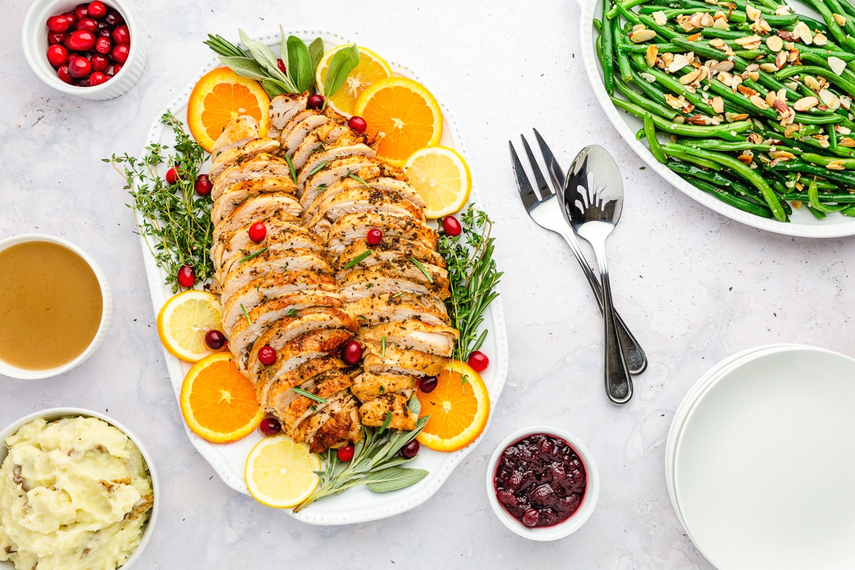 sliced roast turkey breast on a white platter with citrus slices, fresh herbs, and cranberries as garnish with a fork and spoon to the right and cranberry sauce in a bowl to the bottom right.