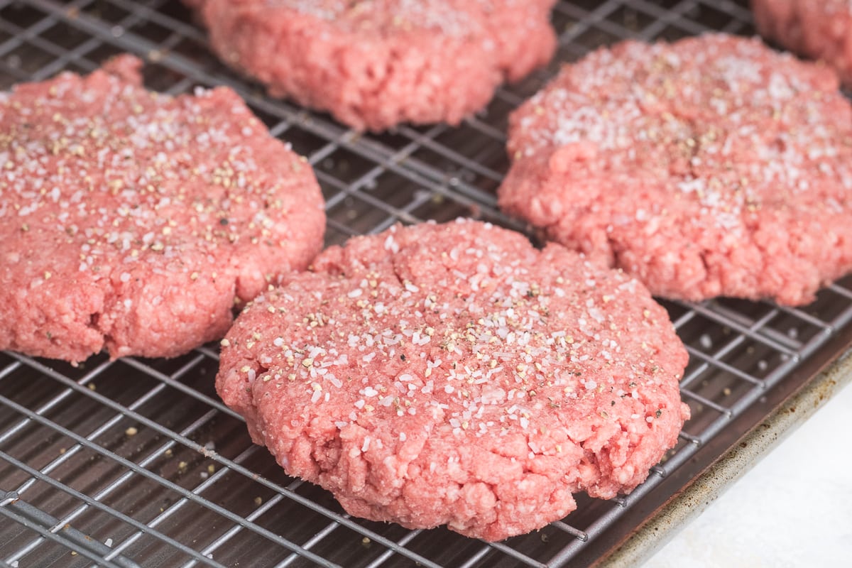 raw hamburger patty on a wire rack ready to be baked with salt and pepper seasoning.