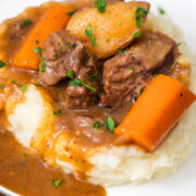 Beef stew with tender beef chunks, carrots, and potatoes served over creamy mashed potatoes, topped with rich brown gravy and garnished with chopped parsley.