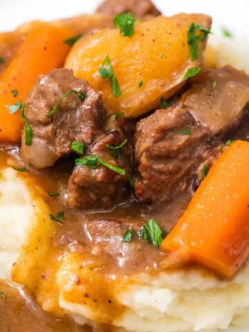 close up of beef stew served over mashed potatoes on a white plate.