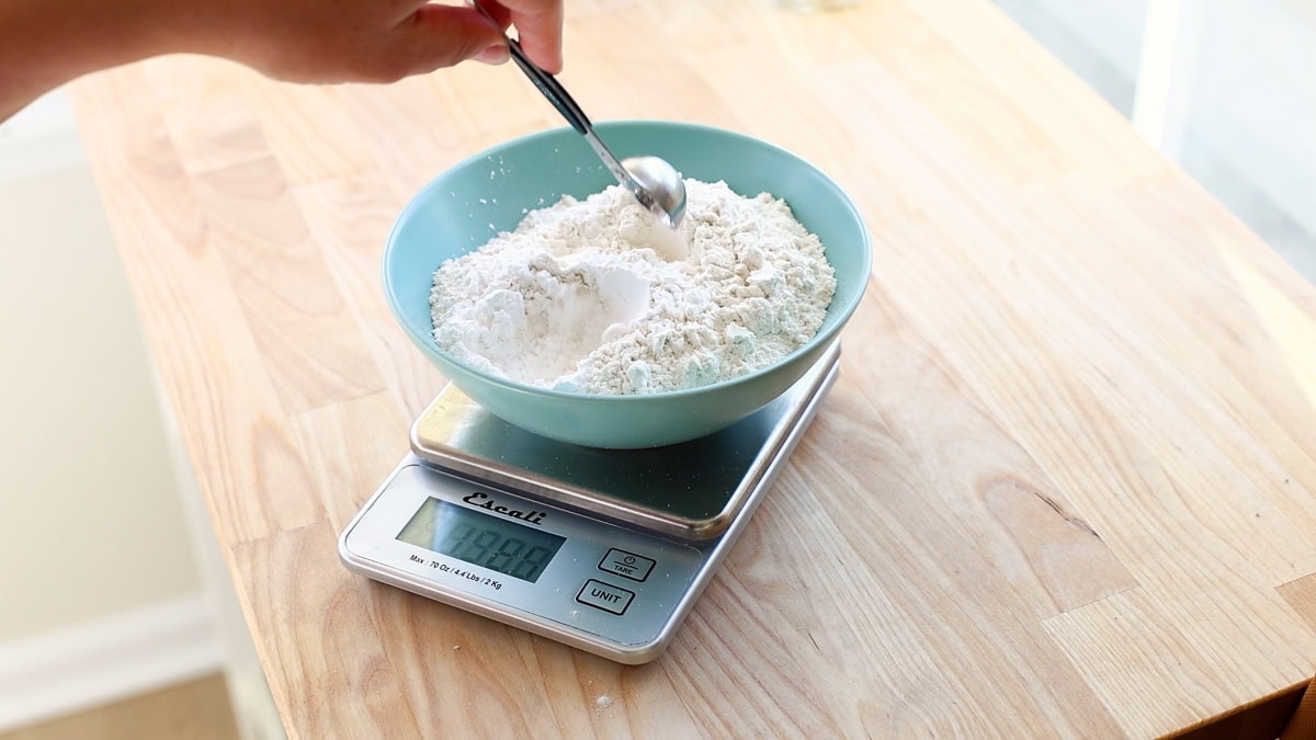 A hand holding a spoon over a bowl of flour and baking powder placed on a digital kitchen scale, preparing to level or add ingredients.