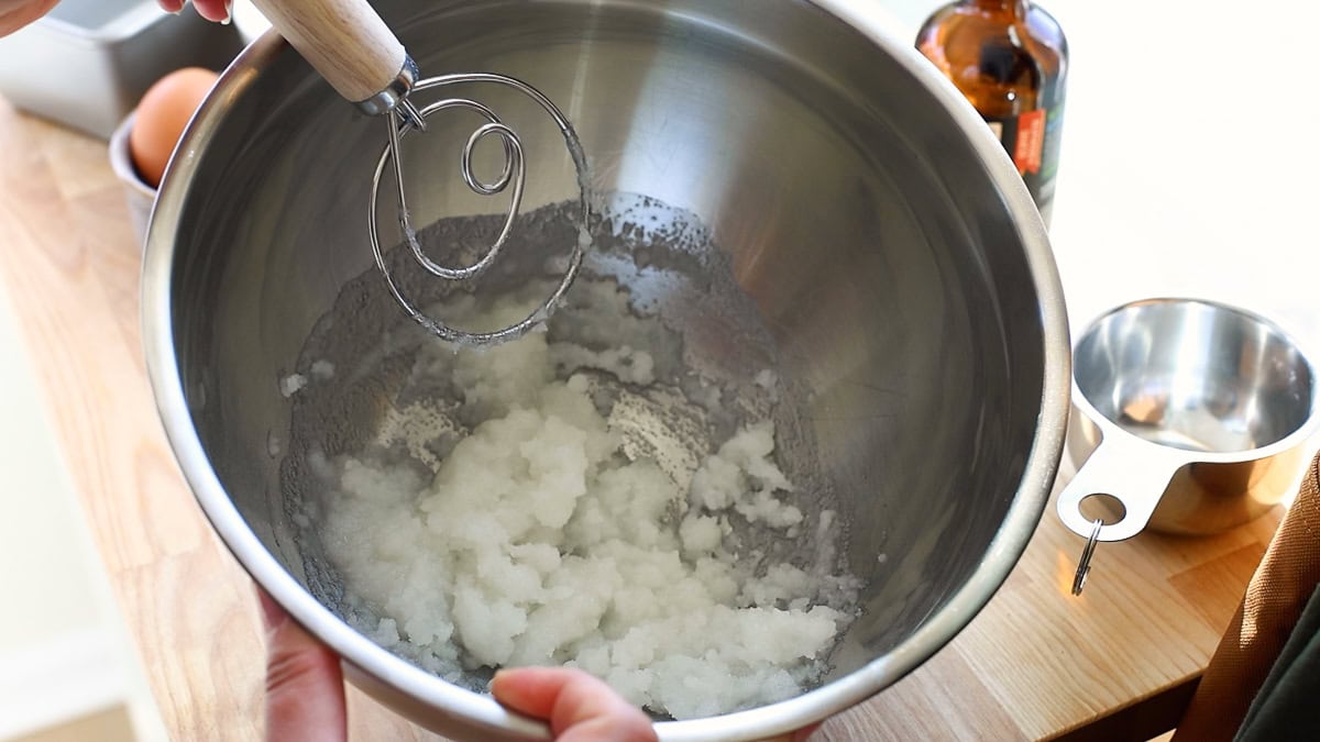 A hand holding a dough whisk over a bowl of creamed sugar and oil, with a bottle of vanilla extract and a measuring cup nearby.