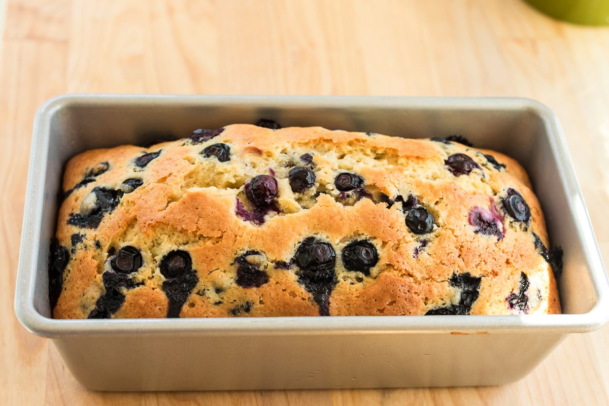 Freshly baked blueberry bread in a loaf pan with golden crust and visible blueberries on top.
