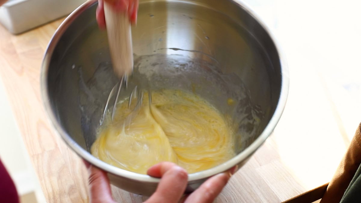 Wet ingredients including eggs being whisked vigorously in a stainless steel bowl.