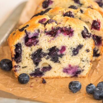 Close up photo of blueberry bread sliced on a piece of parchment paper with fresh blueberries next to it.