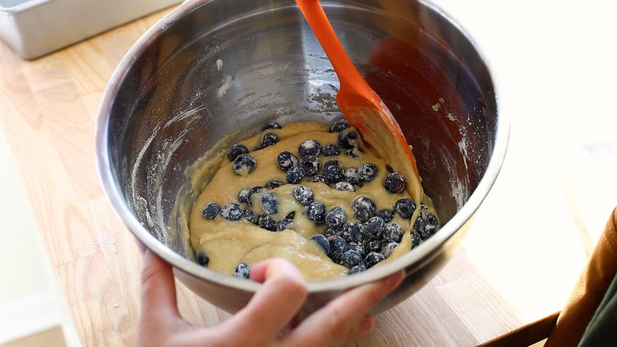 A close-up of the mixing bowl showing flour-coated blueberries being folded into the batter with a spatula.