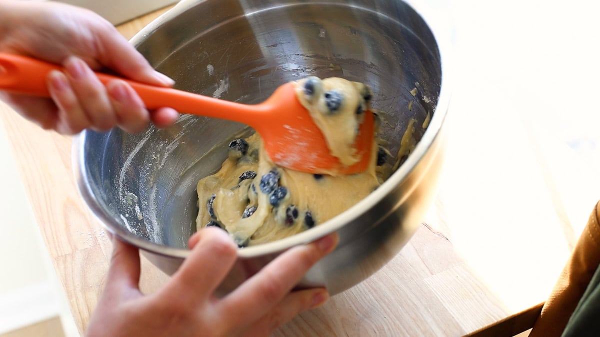 A person using an orange spatula to fold fresh blueberries into thick batter in a large metal mixing bowl.