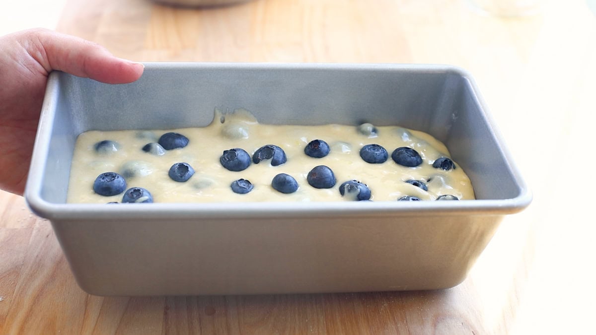 A hand holding a loaf pan filled with thick blueberry bread batter, with blueberries visible on the surface.