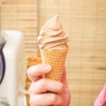 Chocolate soft serve ice cream in a waffle cone held by hand in front of the GreenPan Frost machine