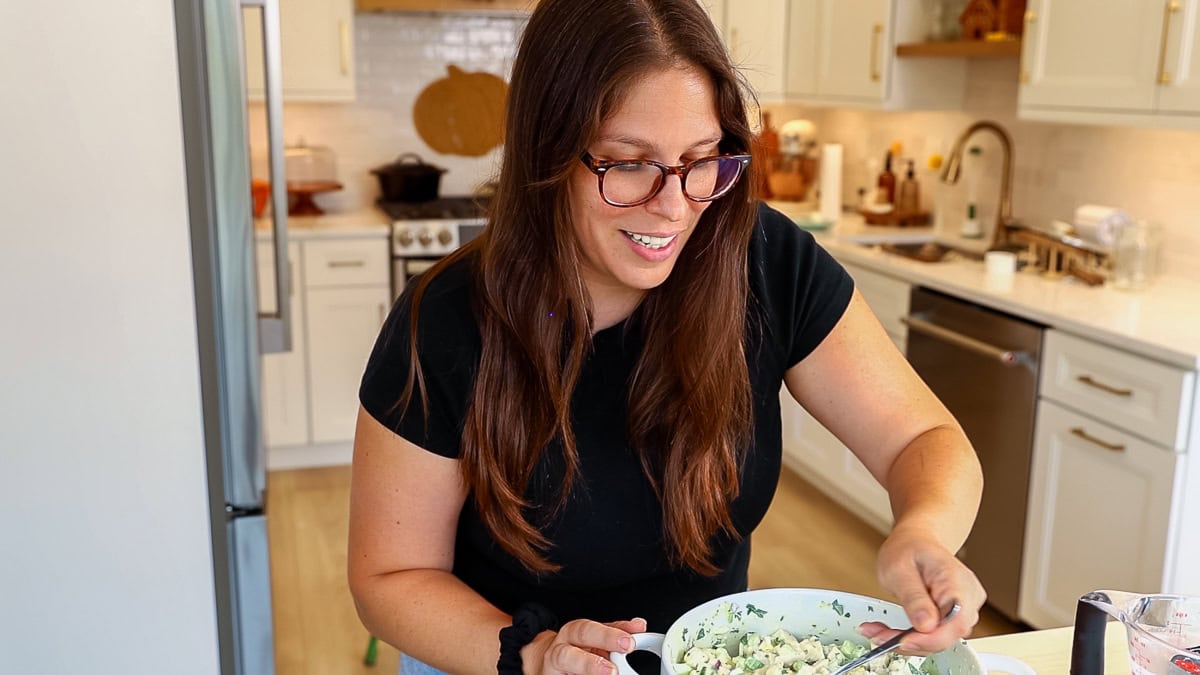 Smiling woman in a black shirt mixing chicken and egg salad in a bright kitchen.