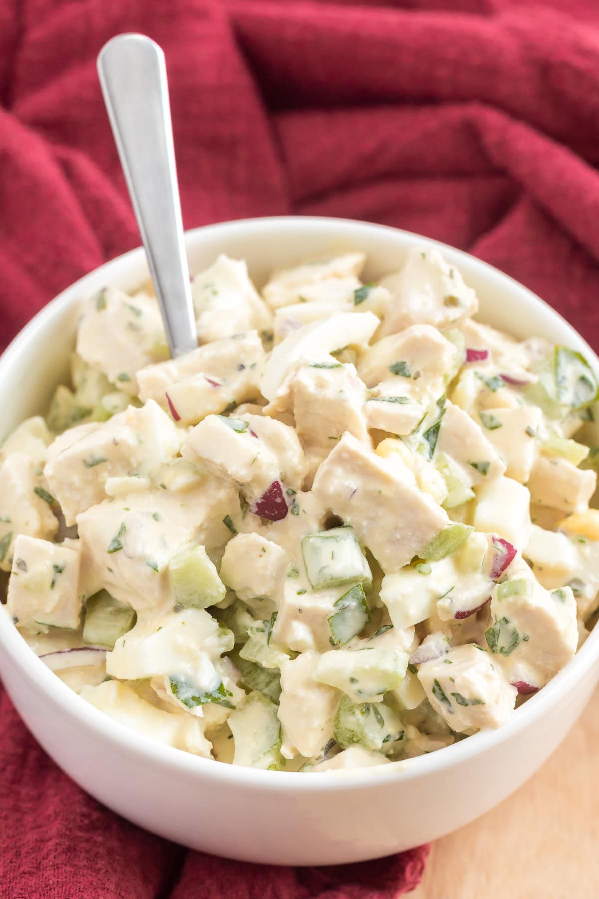 Mixed salad of chicken, eggs, celery, and herbs, with red onion pieces, with a red cloth background.