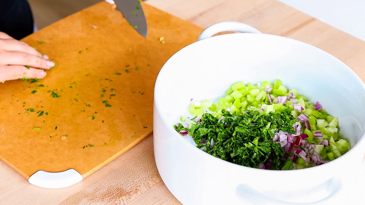 Chopping fresh parsley on a cutting board with diced celery and red onion in a white mixing bowl.