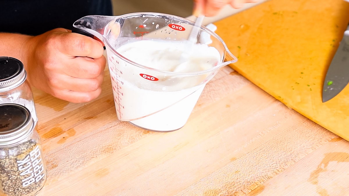 Stirring creamy mayo dressing in a measuring cup with jars of black pepper and salt nearby.