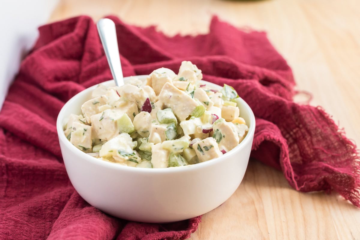 White bowl filled with chunky chicken and egg salad on a wooden surface with a red cloth.