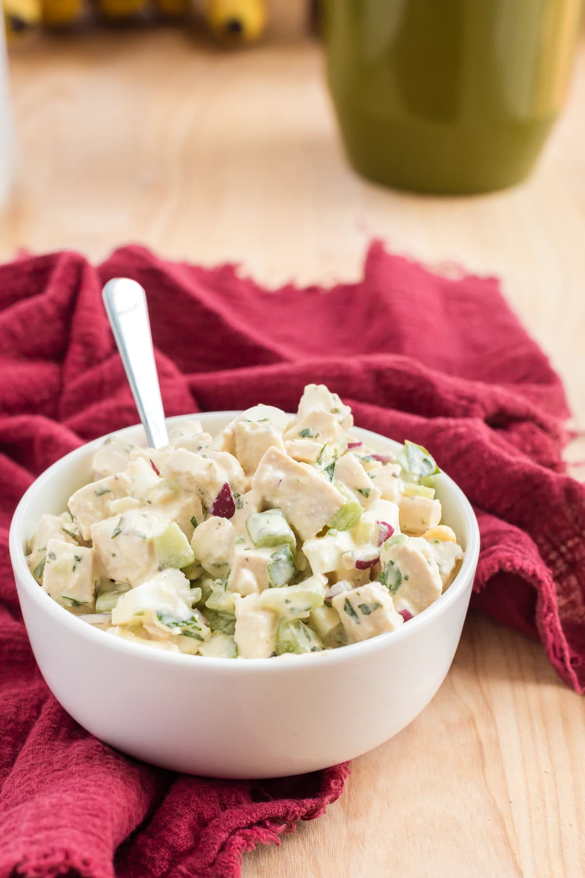 White bowl of creamy chicken and egg salad with celery and herbs, red cloth and blurred kitchen items in background.