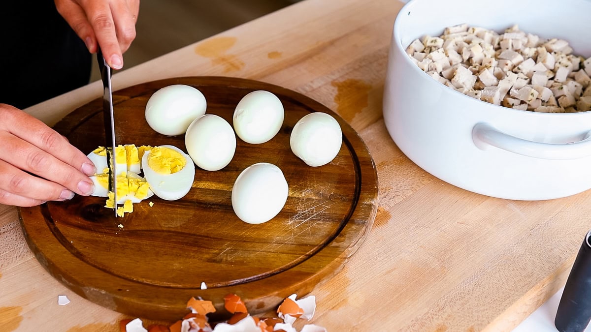 Slicing hard-boiled eggs on a wooden cutting board with chopped chicken in a white mixing bowl nearby.