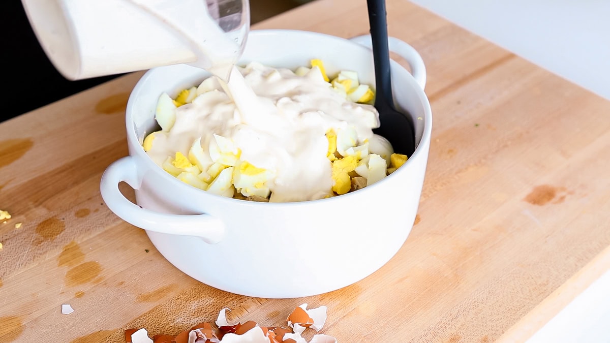 Pouring creamy mayo dressing into a white mixing bowl filled with chopped hard-boiled eggs.