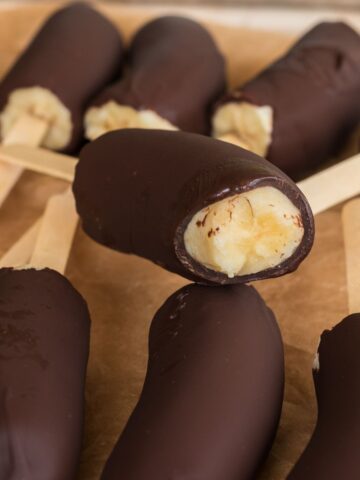 Close up of frozen chocolate covered bananas on popsicle sticks with one showing a bite revealing the creamy banana inside, arranged on brown parchment paper.