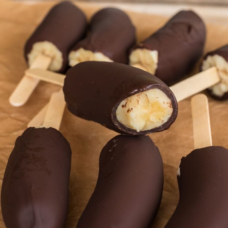 Close up of frozen chocolate covered bananas on popsicle sticks with one showing a bite revealing the creamy banana inside, arranged on brown parchment paper.
