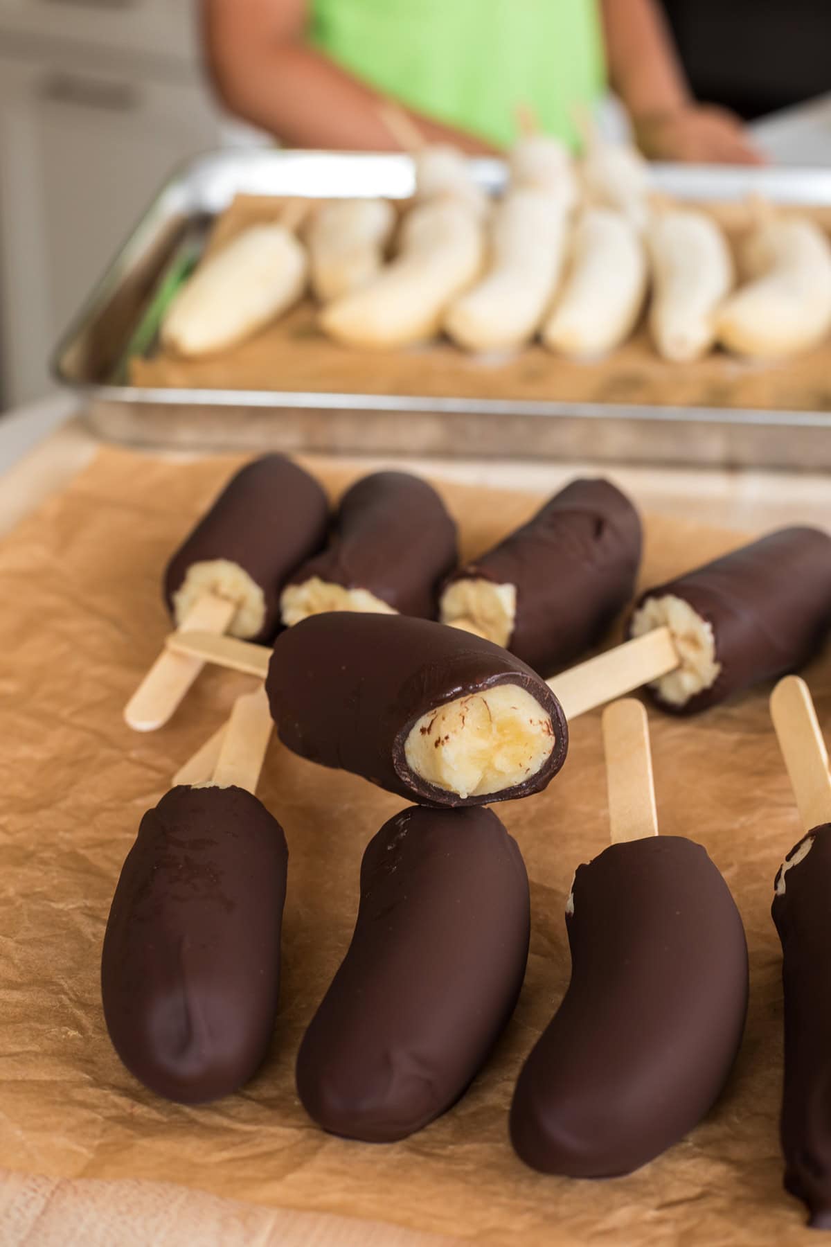 Chocolate-covered banana pops arranged on parchment paper, with frozen undipped bananas on a tray in the background.
