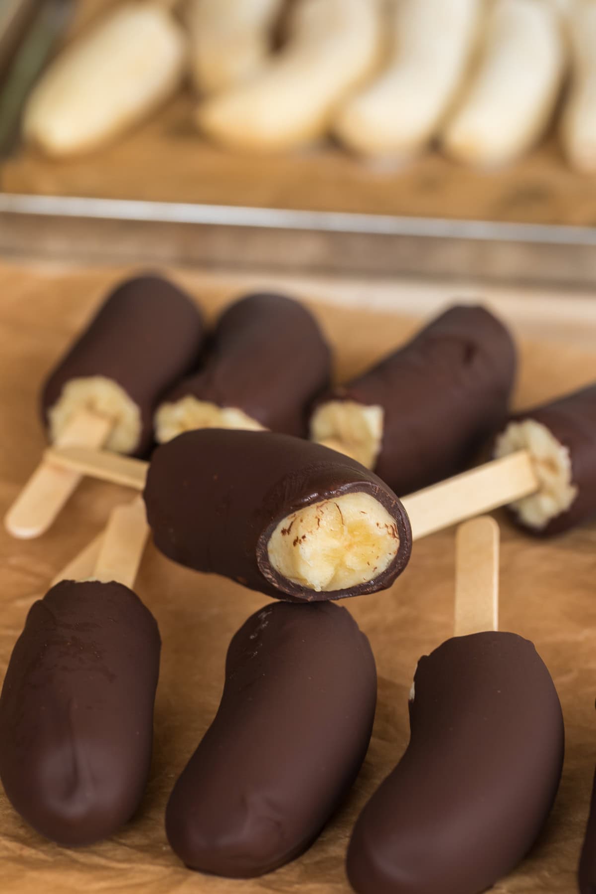 Close-up of chocolate-covered banana pops on sticks, one with a bite taken out to reveal the creamy banana inside, with undipped bananas blurred in the background.