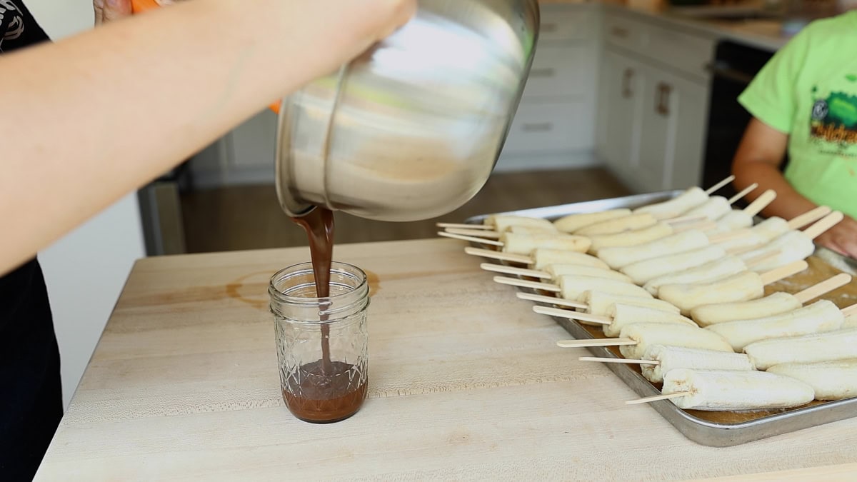 Pouring melted chocolate from a saucepan into a glass jar, with a tray of frozen bananas on sticks in the background.