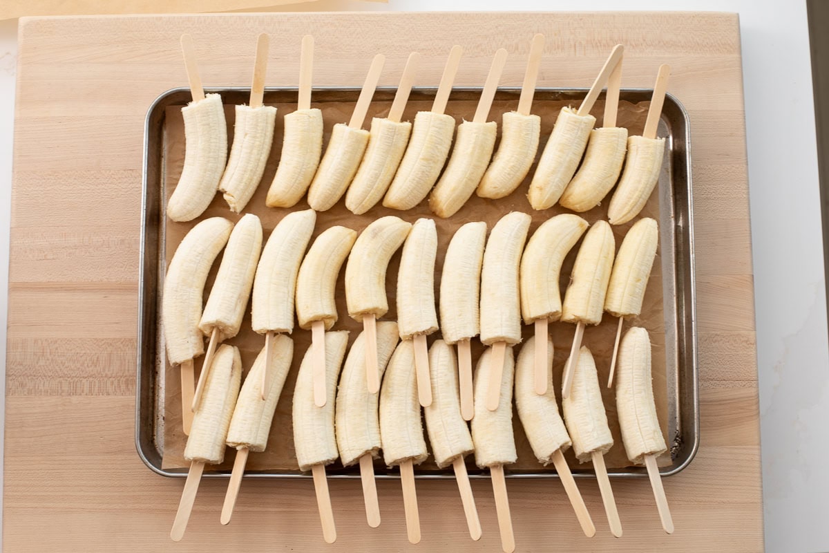 Peeled banana halves with wooden popsicle sticks inserted into one end, arranged on a baking sheet.