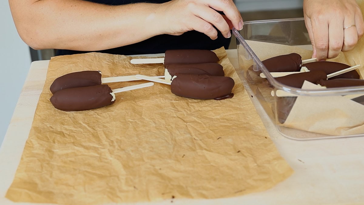 Placing finished chocolate-covered banana pops into a freezer safe container lined with parchment paper to place in the freezer.