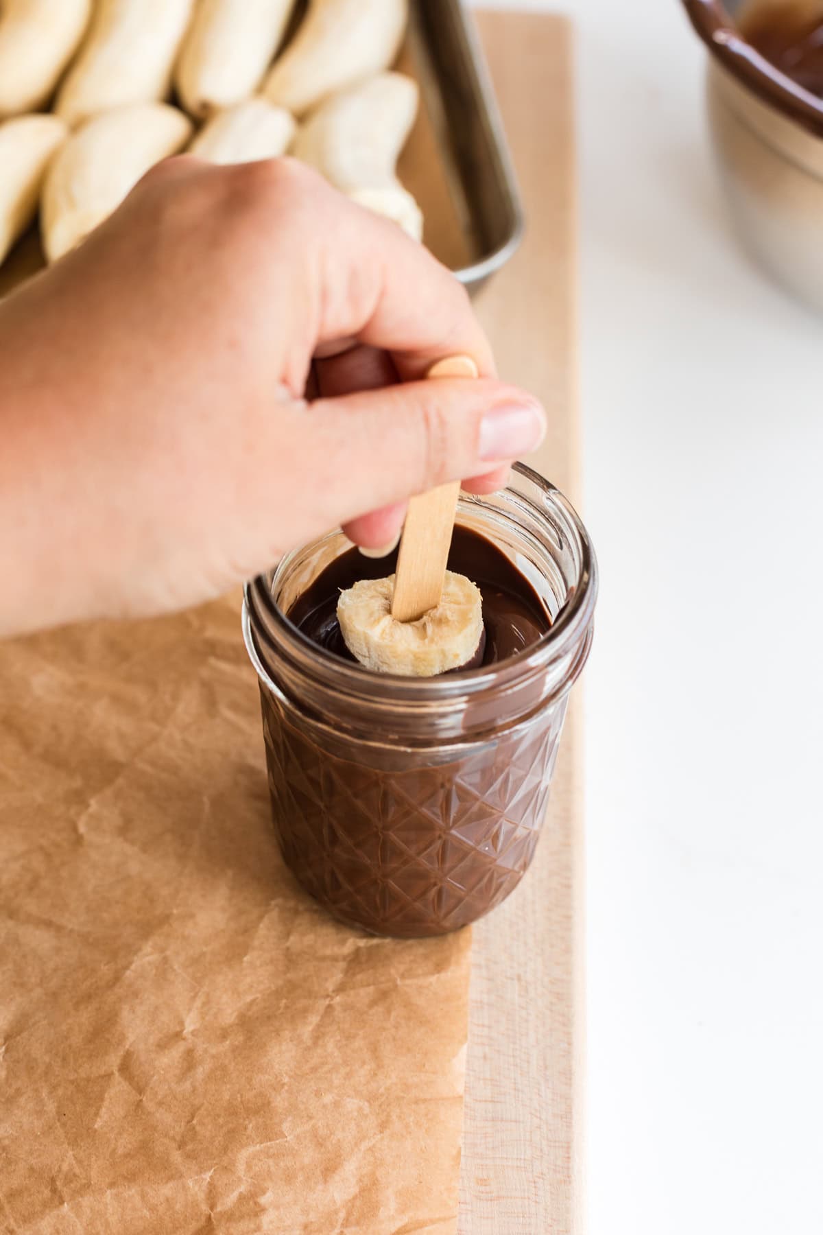 Hand dipping a banana half on a stick into a jar of melted chocolate.