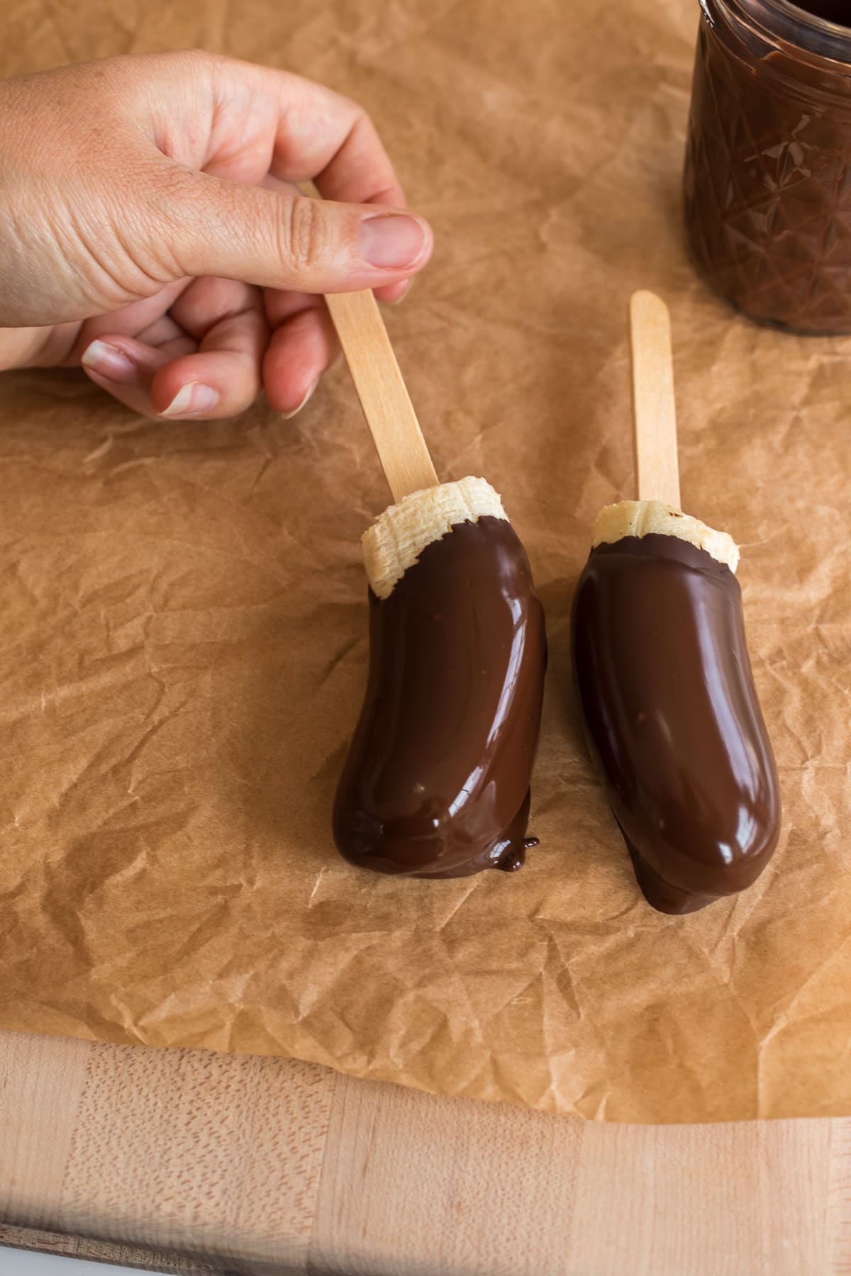Two chocolate-dipped banana halves on sticks resting on brown parchment paper.