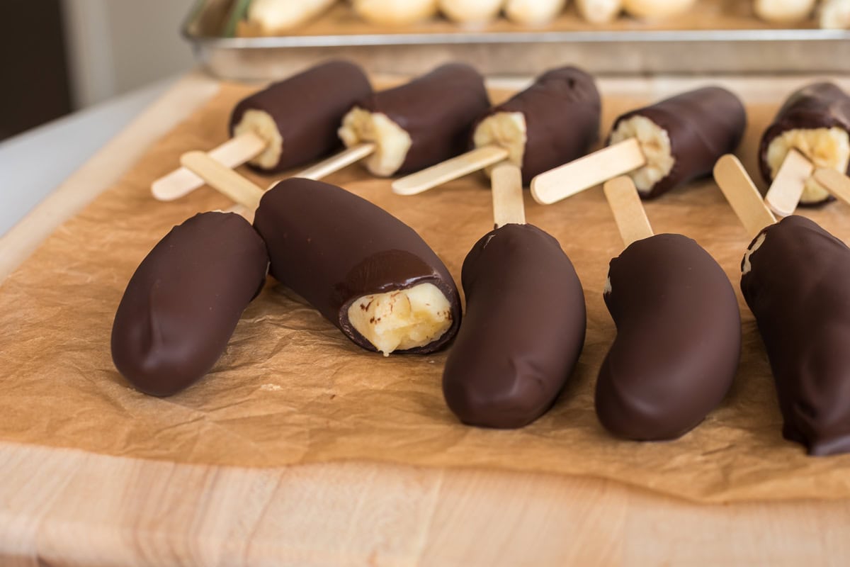 Several chocolate-covered banana pops on parchment paper, one with a bite taken to reveal the banana inside.