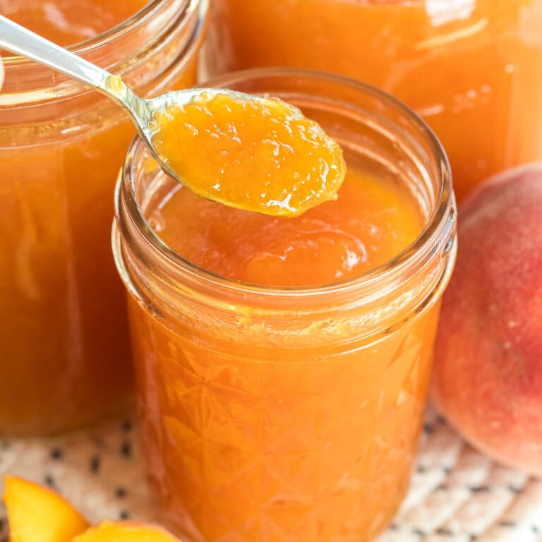 Close up spoon scooping peach jam from a glass jar with fresh peaches beside it.