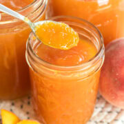 Close-up of a spoonful of peach jam above a jar, surrounded by jars of jam and peaches.