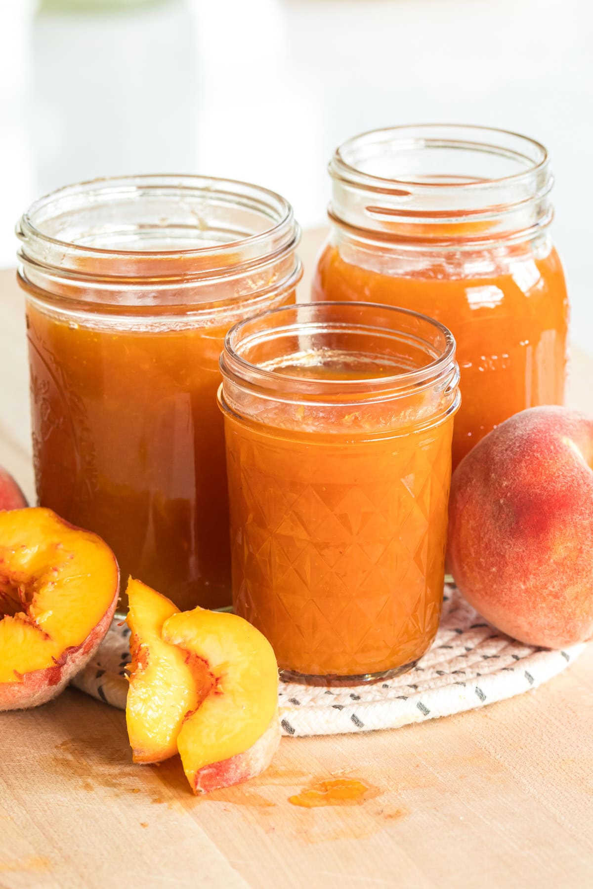 Homemade peach jam in three jars with fresh peaches and peach slices arranged in front.