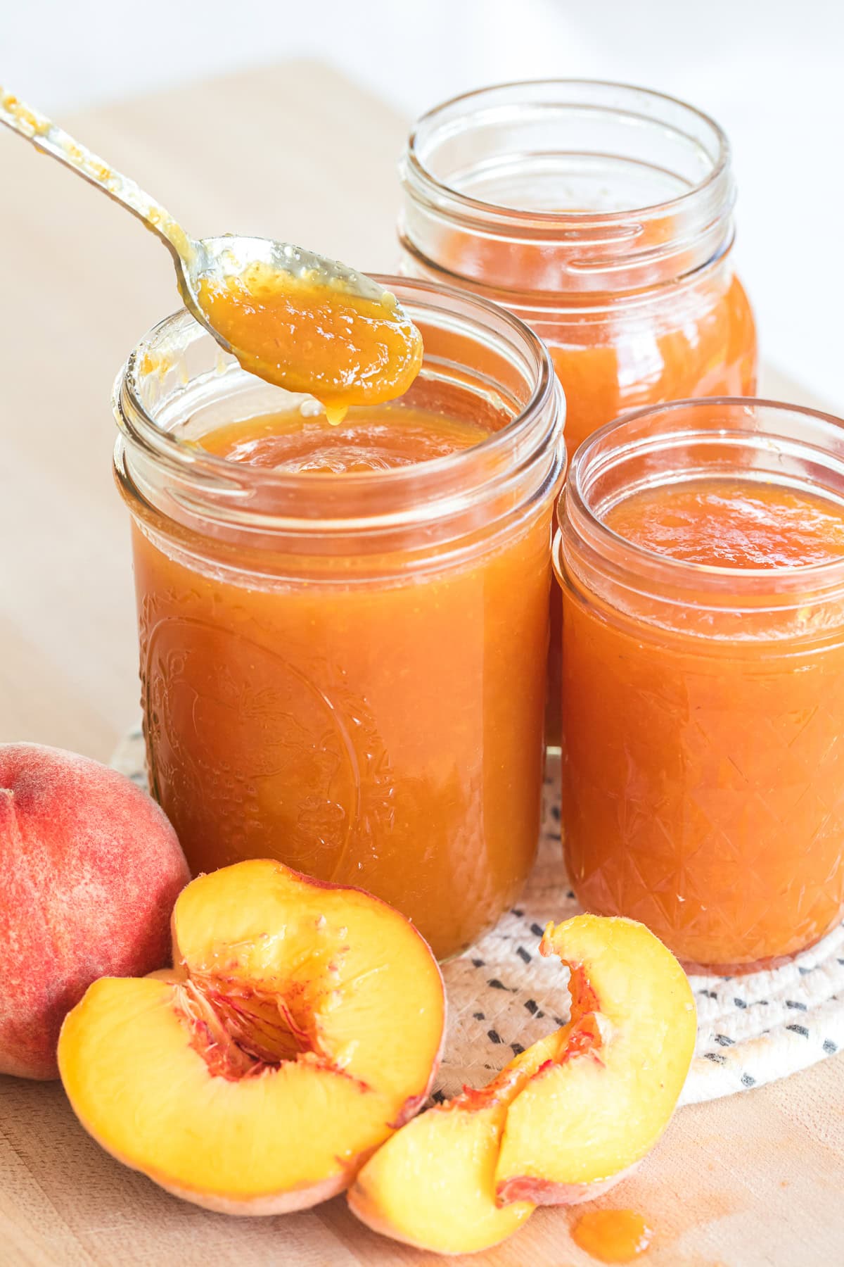 Spoon scooping peach jam from a glass jar with fresh peaches beside it.