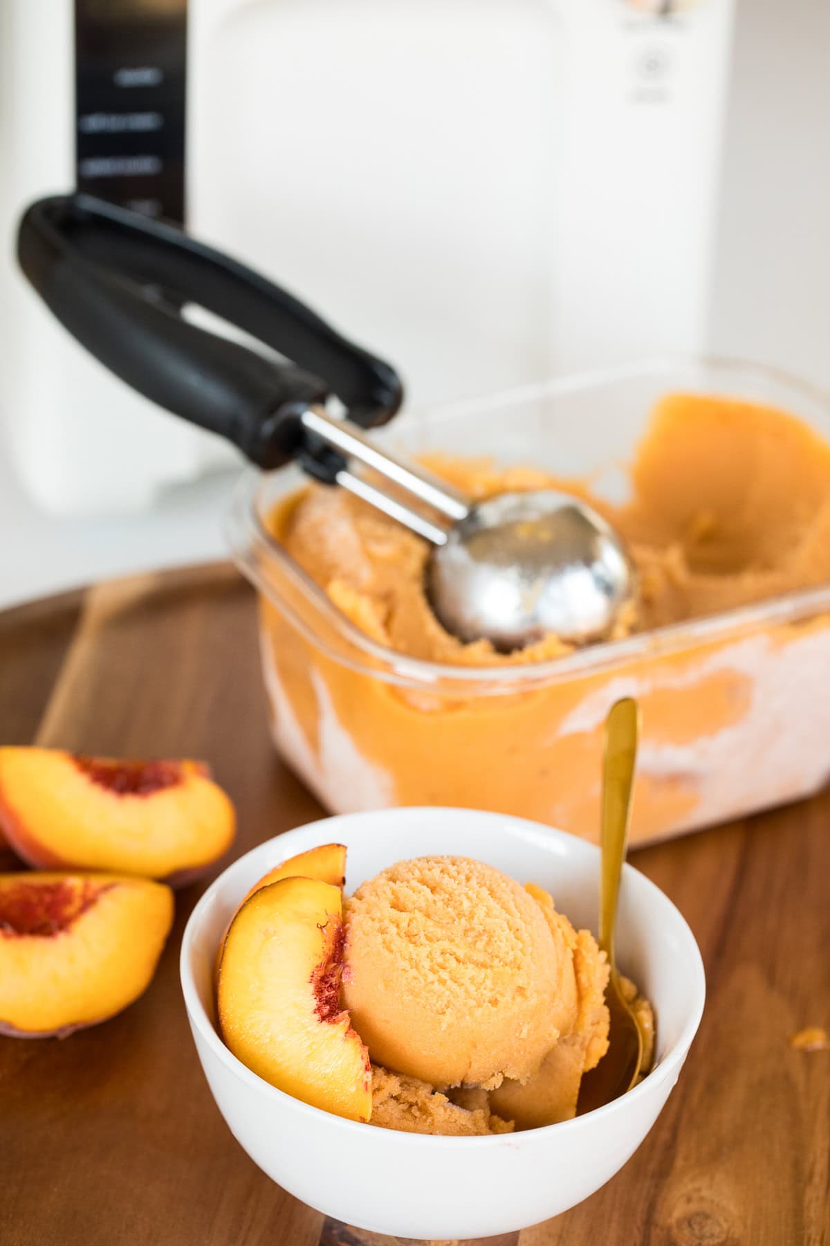 A serving bowl of peach sorbet with peach slices and a gold spoon beside the container of sorbet in front of the GreenPan frost ice cream and frozen treat maker.