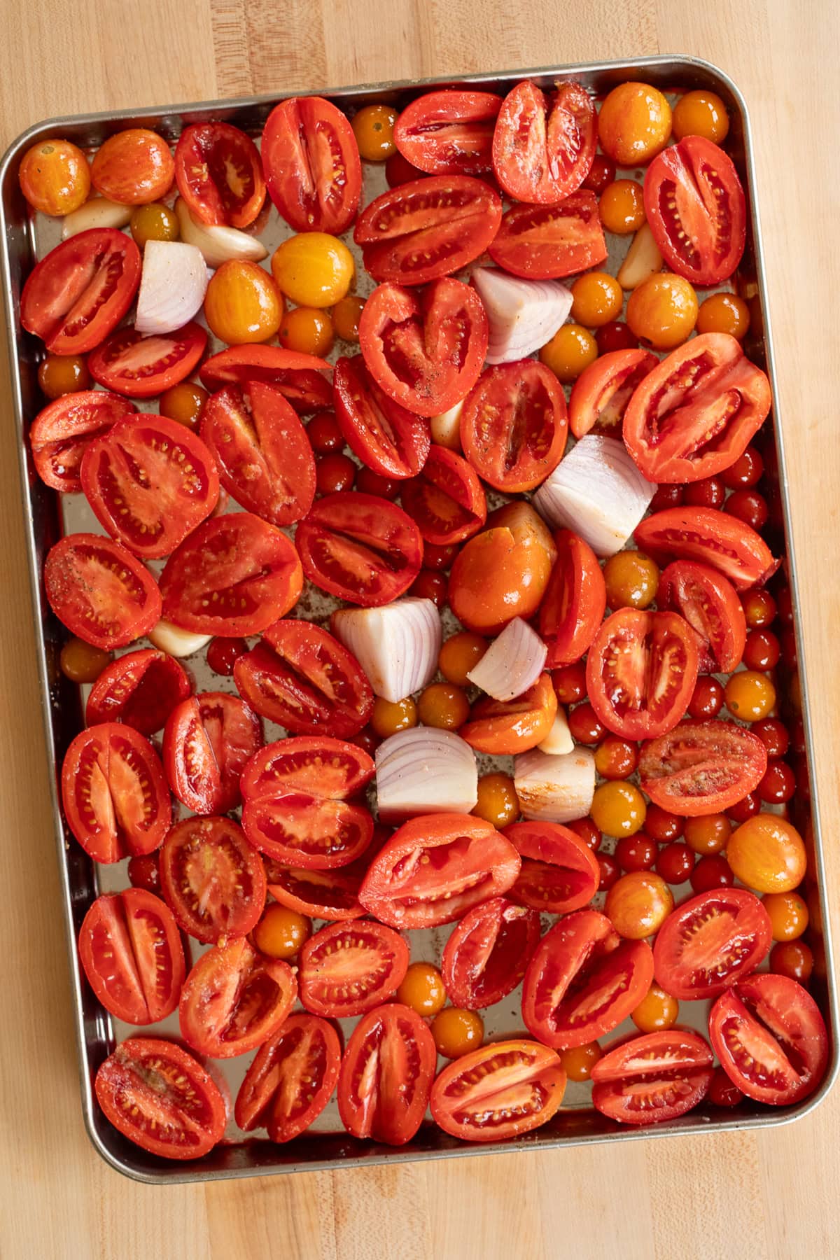 Overhead view of halved tomatoes, cherry tomatoes, and quartered onions spread on a baking sheet before roasting.