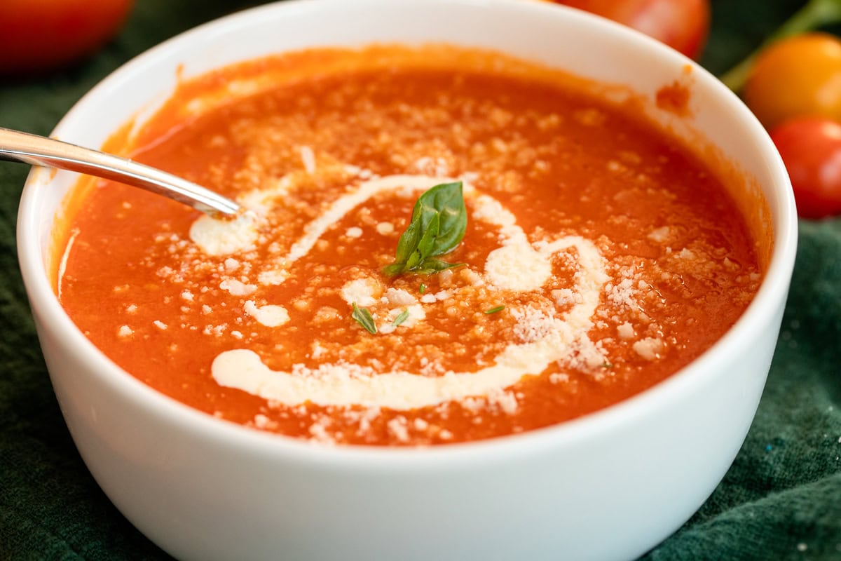 Warm bowl of fresh roasted tomato basil soup with cream swirl and Parmesan topping, served on a green cloth.