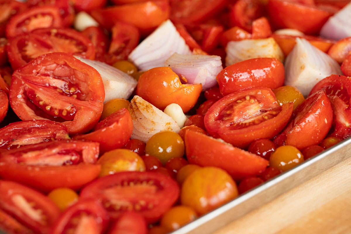 Fresh tomatoes, onions, and garlic tossed with olive oil on a sheet pan, ready for roasting.