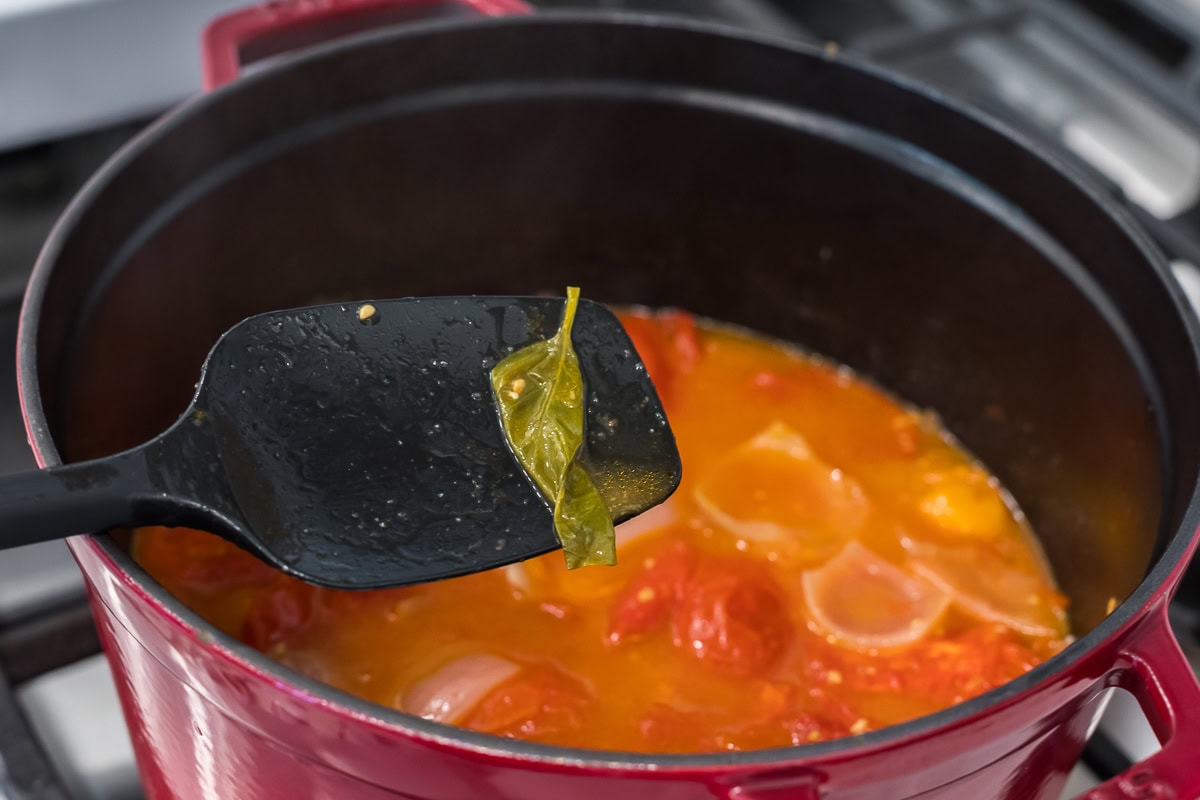 Cook removing a basil leaf from simmering roasted tomato soup with a black spatula.