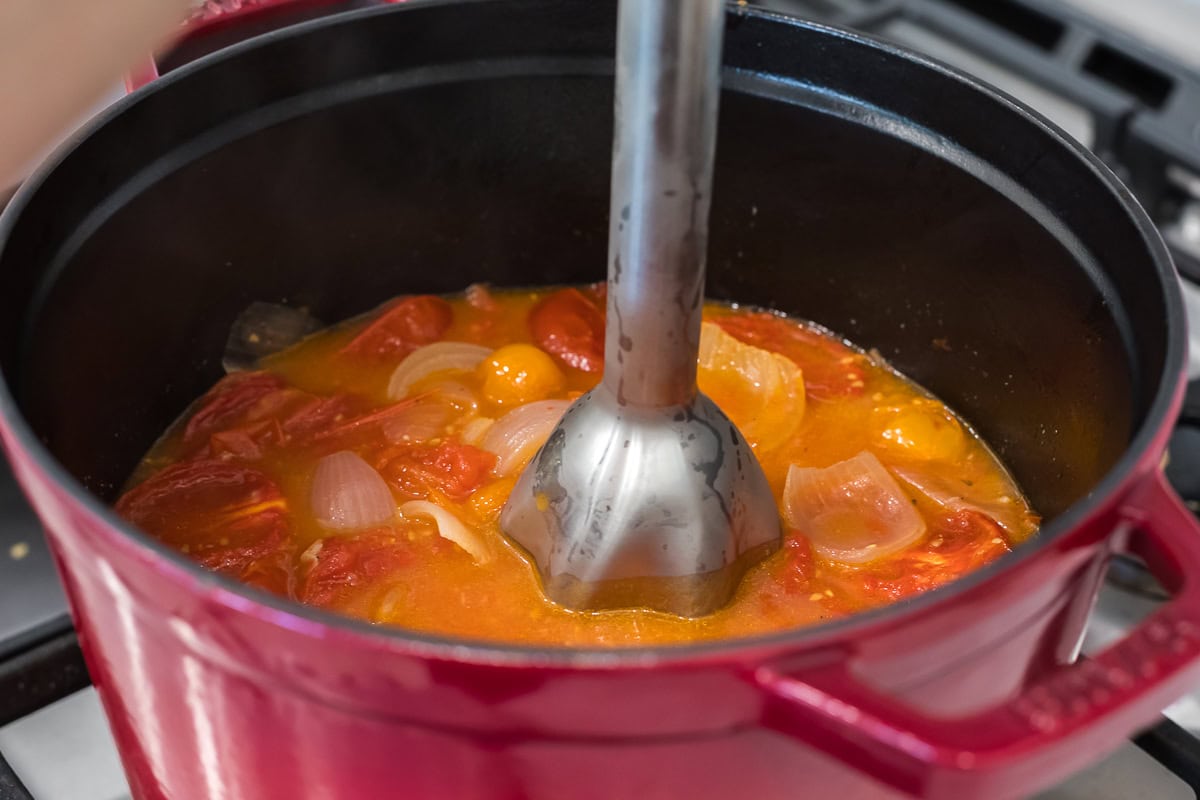 Immersion blender pureeing roasted tomatoes, onions, and garlic in a red Dutch oven to create a creamy soup base.