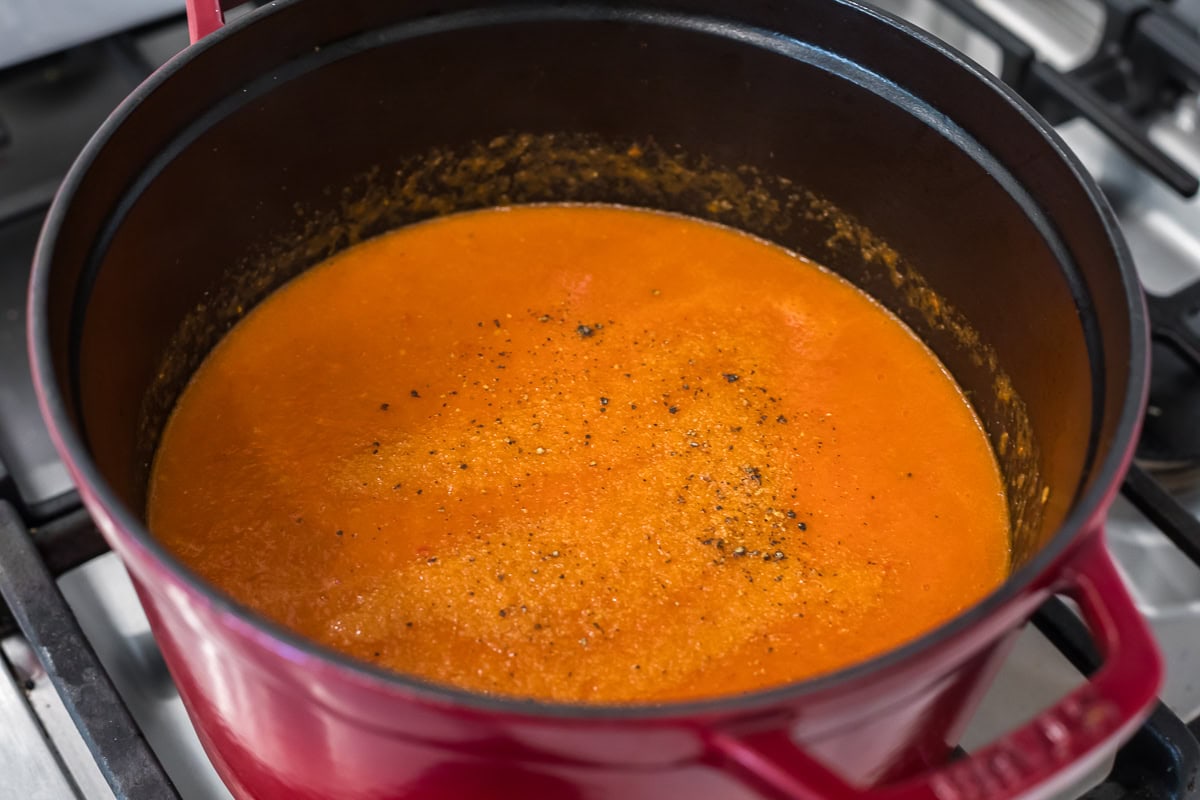 Smooth roasted tomato soup simmering in a red Dutch oven, speckled with freshly cracked black pepper, granulated garlic, and granulated onion.