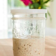 A glass Ball mason jar with a white plastic lid, filled halfway with bubbly sourdough starter, sits on a wooden countertop. In the blurred background, a bouquet of vibrant flowers adds a pop of color in a bright kitchen.