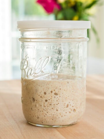 A glass Ball mason jar with a white plastic lid, filled halfway with active bubbly sourdough starter, sits on a wooden countertop. In the blurred background, a bouquet of vibrant flowers adds a pop of color in a bright kitchen.