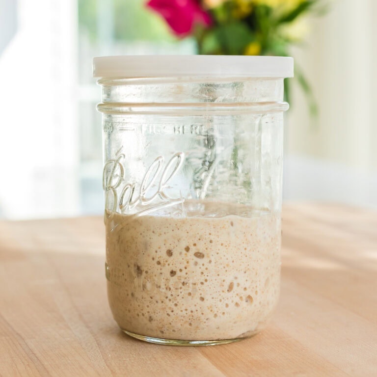 A glass Ball mason jar with a white plastic lid, filled halfway with active bubbly sourdough starter, sits on a wooden countertop. In the blurred background, a bouquet of vibrant flowers adds a pop of color in a bright kitchen.